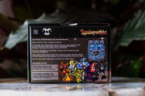 The back of the Ignisynthia box showing text and imagery from the game, standing upright on a reflective surface with a blurred dark green garden background.