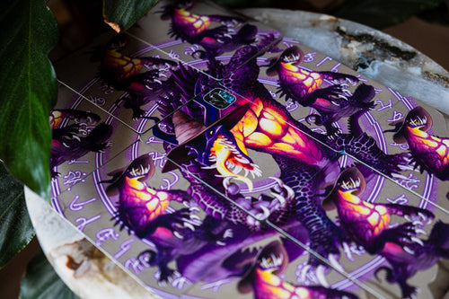 A close-up of cards forming a spell circling with detailed fantasy illustration of a purple dragon with glowing orange and yellow eyes, surrounded by occult-style symbols on a light background. Dark green leaves partially frame the left side of the image.