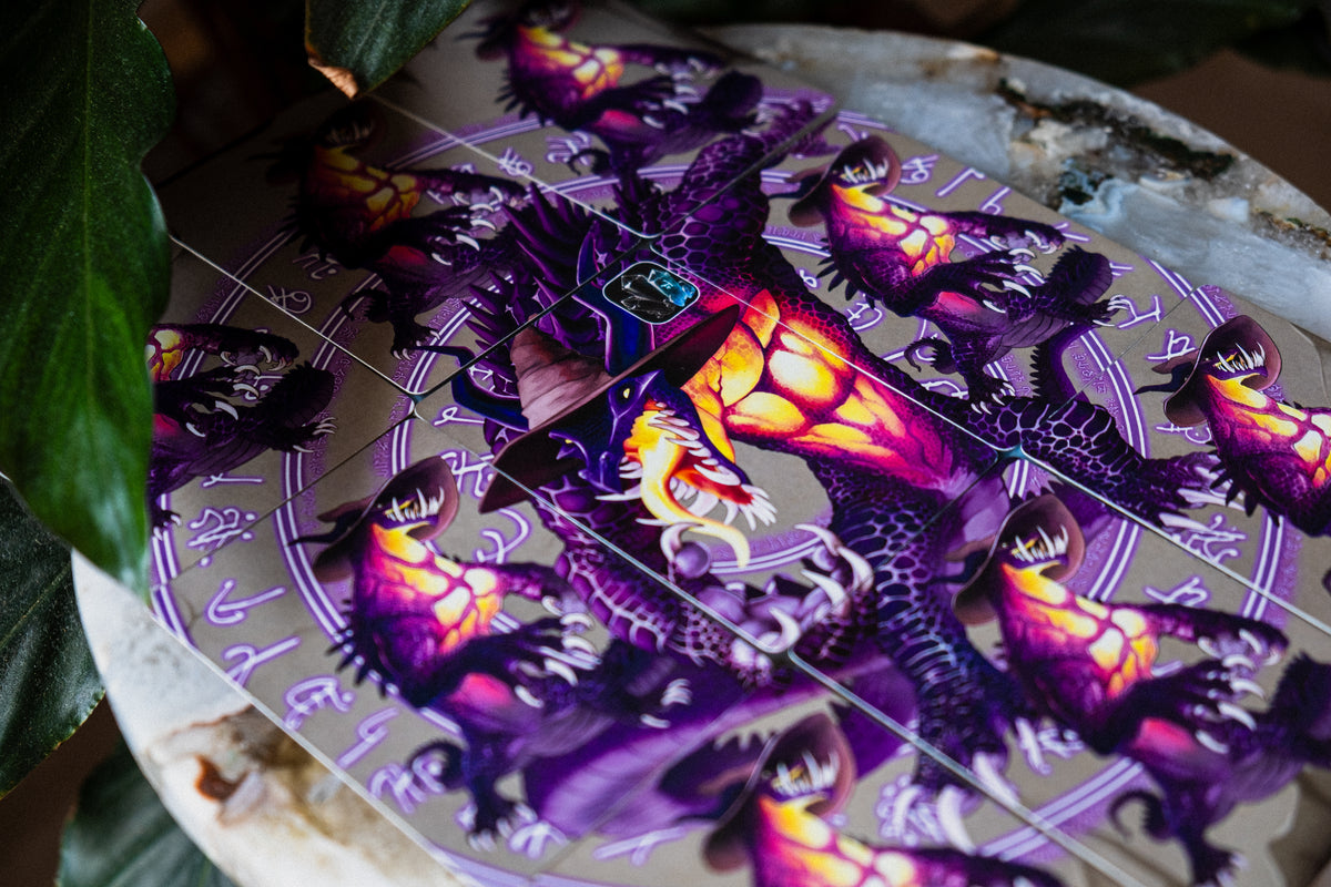 A close-up of cards forming a spell circling with detailed fantasy illustration of a purple dragon with glowing orange and yellow eyes, surrounded by occult-style symbols on a light background. Dark green leaves partially frame the left side of the image.