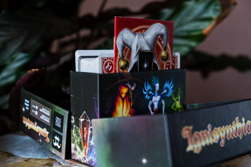 A tabletop close-up of a boxed card game set with illustrated packaging. The front box is dark with fantasy artwork and the visible title text reads 'Ignisynthia'.