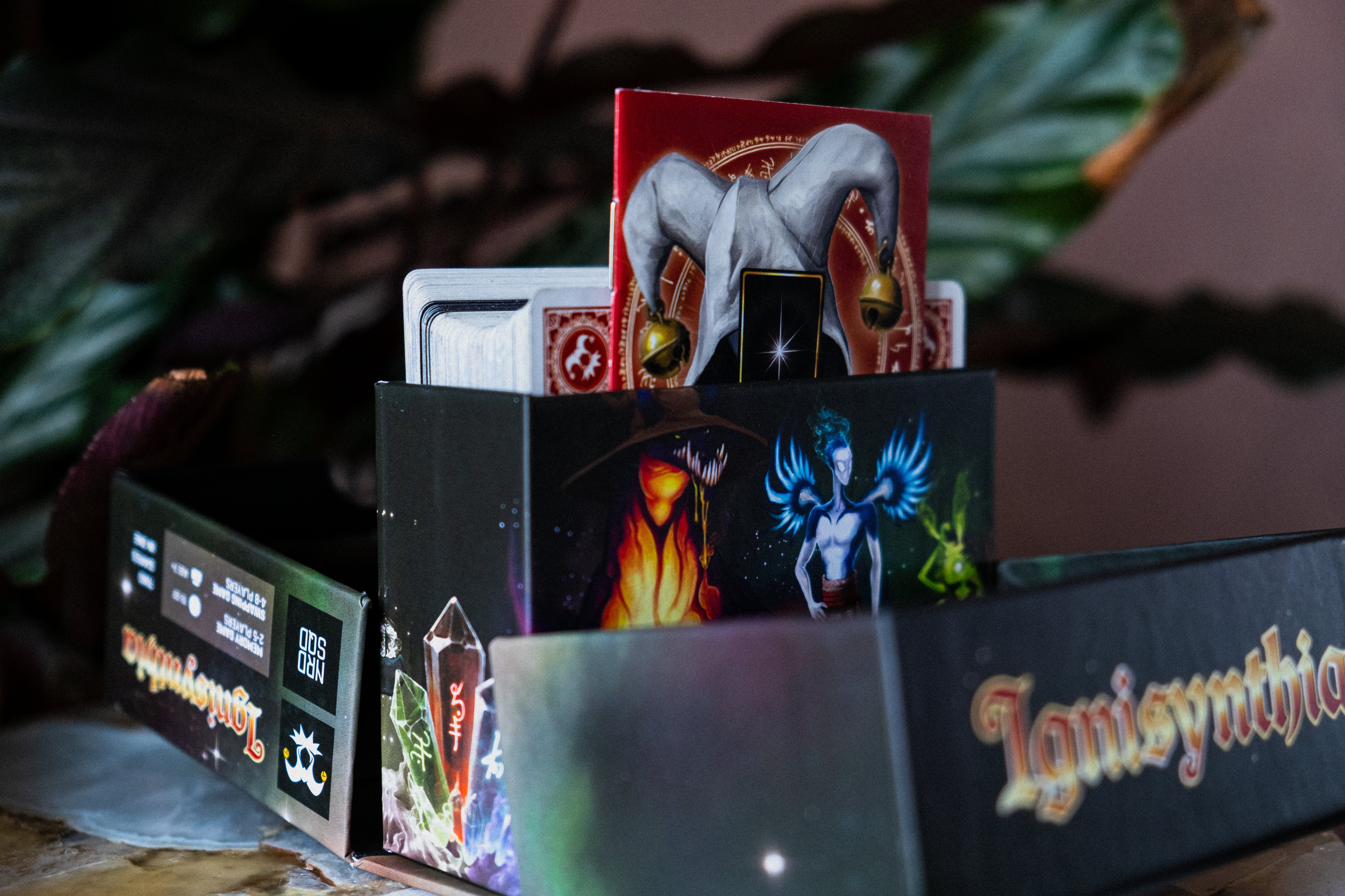 A tabletop close-up of a boxed card game set with illustrated packaging. The front box is dark with fantasy artwork and the visible title text reads 'Ignisynthia'.